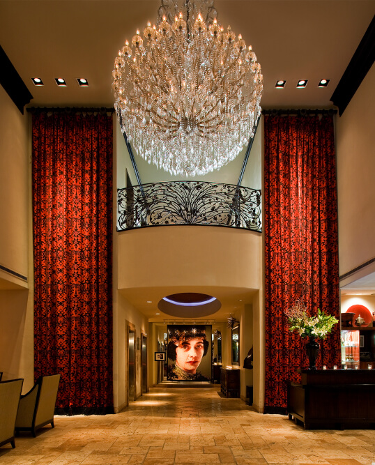 Grand hotel lobby with giant crystal chandelier, red damask drapes, ornate balcony, and a large portrait at the far end.